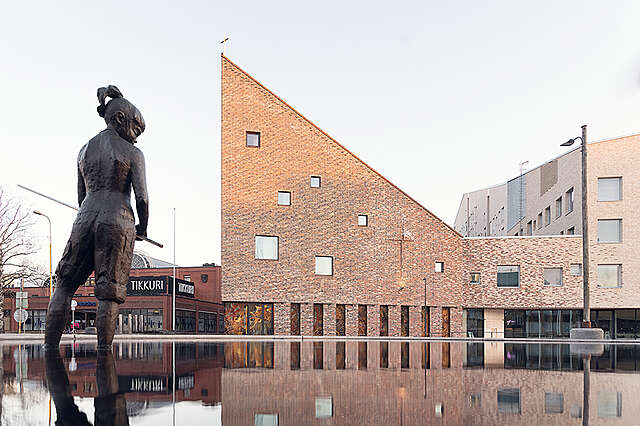 Taustalla punatiilinen, jyrkän viistokattoinen rakennus, joka peilautuu vesialtaasta. Etualalla pronssinen patsas seisoo altaassa.