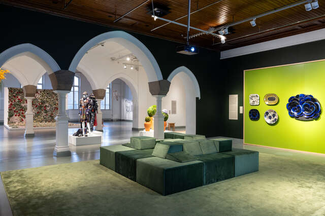 Näyttelytilan seinillä taidekeramiikkaa ja keskellä nelionmuotoinen sohvaryhmä, tilan holvikaarien takana puettuja mannekiineja ja kasvitaulu