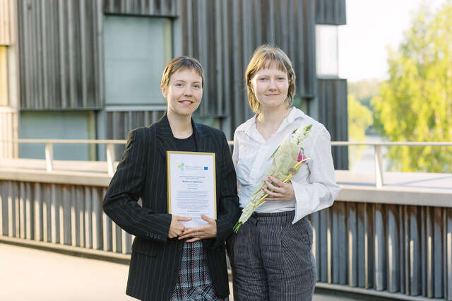 Kaksi nuorta naista, toisella kädessä palkinto ja toisella kukkakimppu.
