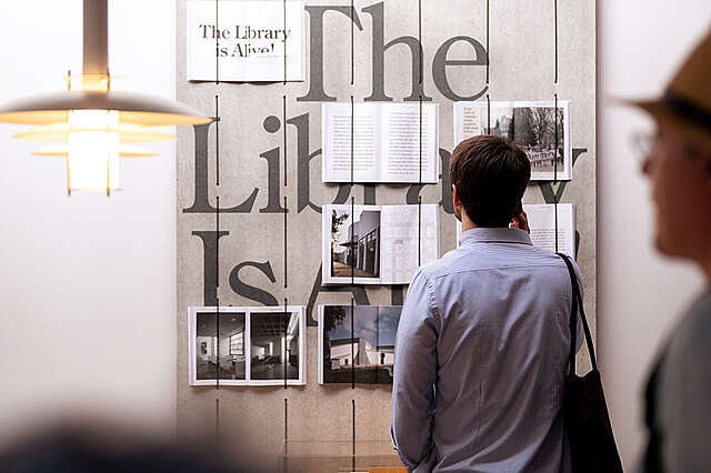 Mind-building näyttely: kaksi henkilöä katsomassa näyttelyn esillepanoa. Seinälle on nostettu levy josta erottuu sanat "the Library is..". Levyn päälle on vaijereilla avattu kirjoja.