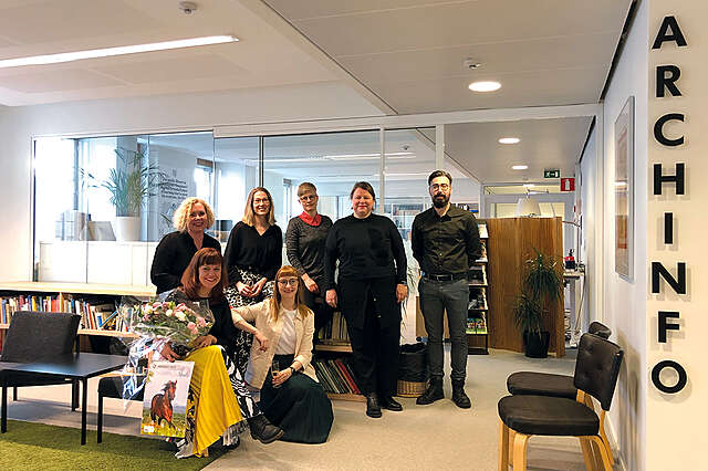 Archinfon seitsemän työntekijää toimiston aulassa iloisessa ryhmäkuvassa.