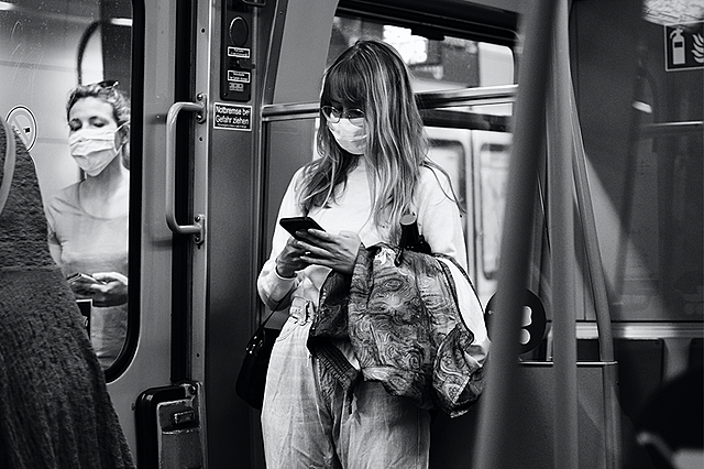 Nainen selaa puhelinta metrossa kasvomaski kasvoillaan.