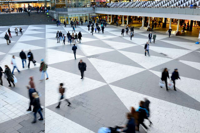A square with triangular colours in the paving seen from above, people walking from place to place.