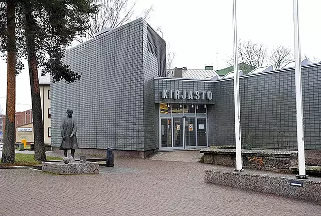 Kirjaston harmaa pääjulkisivu, etualalla vanhaa naista esittävä patsas