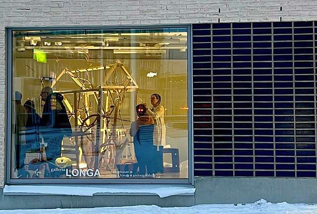 Ulkoa otettu kuva rakennuksesta, jossa suuri ikkuna. Sisällä ihmisiä ja puutaideteos.