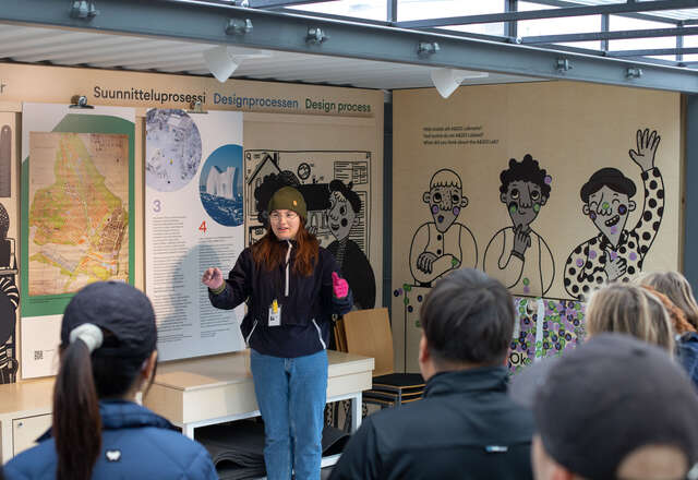 Nainen pipo päässä seisoo etuseinästä avoimessa kontissa ja puhuu yleisölle. Taustaseinällä piirroksia ja muuta näyttelyaineistoa.