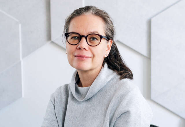 Silmälasipäinen nainen harmaassa neulepaidassa katsoo kameraan.