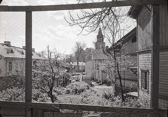 Mustavalkovalokuva, otettu puutalon ikkunan takaa näkyvästä sen edessä näkyvästä puutarhasta.