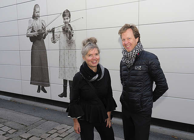 Pariskunta mustissa vaatteissa, taustalla valkoinen seinä jossa kuva kahdesta viulistista.