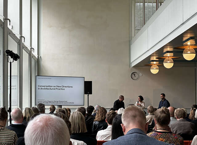 Ihmisiä istuu kuuntelemassa edessä olevia puhujia korkeassa vaaleassa tilassa.