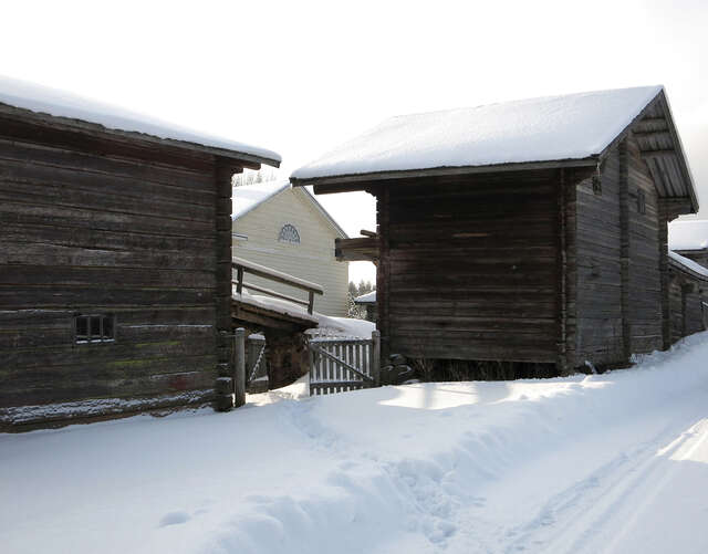 A snowy view from between two dark-coloured log outbuildings towards a white house