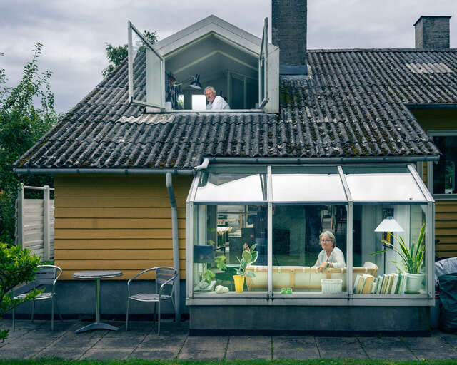 Yellow wooden house, man looks out the upstairs window and woman sits at the patio.