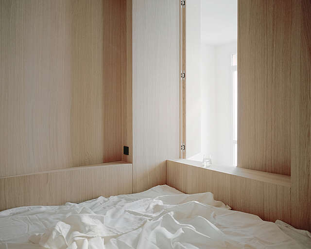 Detail of a room with wooden surfaces, light coming in from an opened hatch, bed covers on the floor