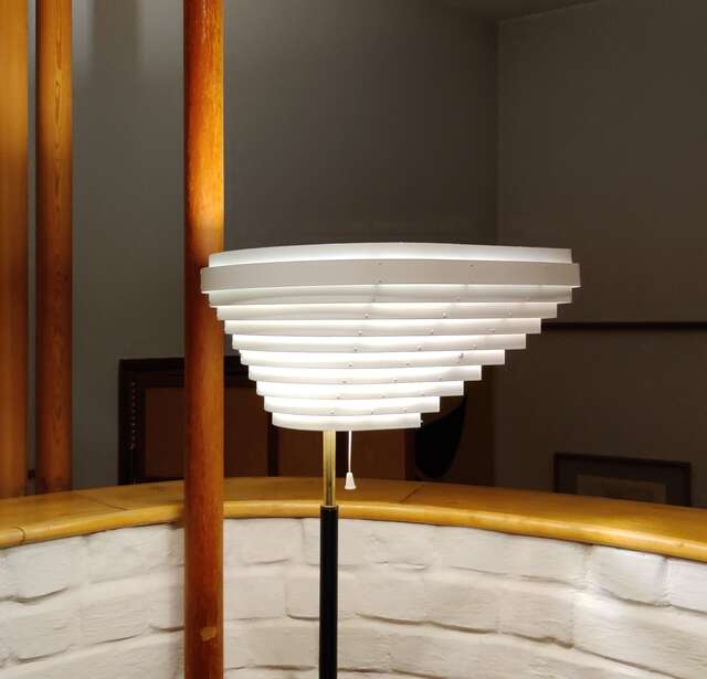 Viuhkamainen, kerroksellinen jalkalamppu, taustalla kaartuva valkoinen tiiliseinämä ja ohuita puupilareita.