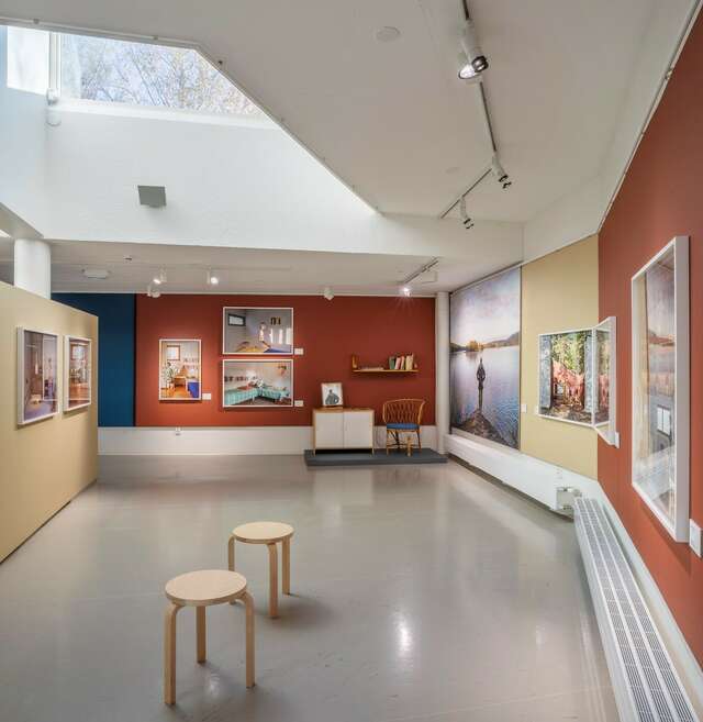 Näyttelytila, jonka seinillä tauluja ja lattialla kaksi pystyssä olevaa puista jakkaraa