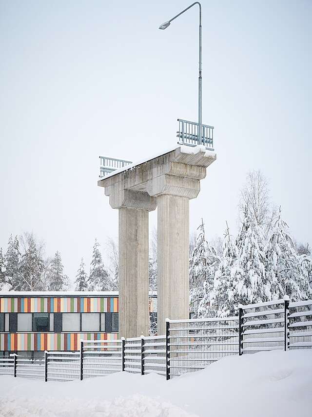 Taideteos, joka muistuttaa betonisesta ajosillasta leikattua viipaletta, jonka huipulla katuvalopylväs ja metalliaidan pätkiä. Takana harmaansininen taivas ja alla lumihanki.