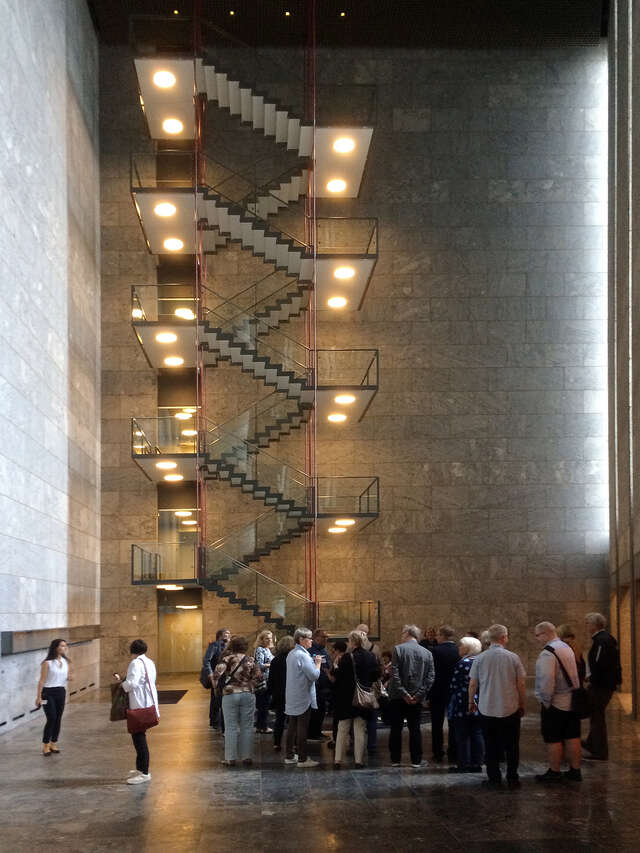 Ryhmä ihmisiä seisoo korkeassa aulatilassa, jonka takaseinustalla valaistu portaikko.