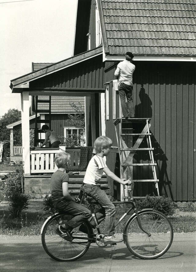 Mustavalkoinen kuva puutalosta, jonka räystästä joku tutkii tikapuilla, edessä pyöräileviä lapsia.