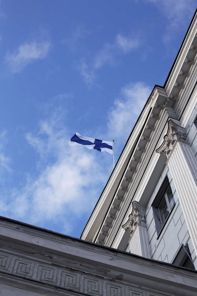 View from bottom to top towards the Finnish flag flying on the roof of the blue-grey classic house. Blue sky, a few wispy clouds.