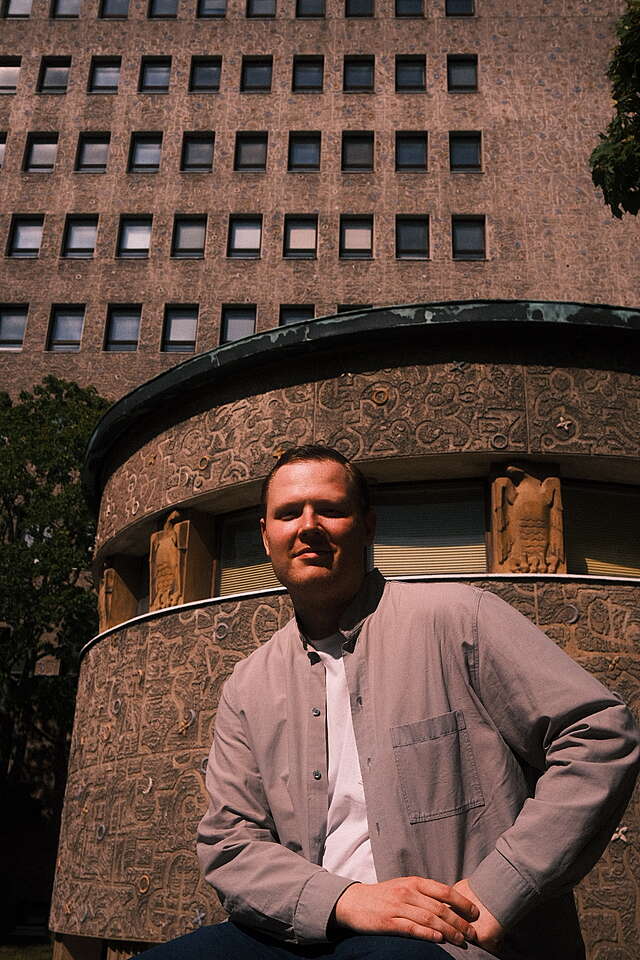 A smiling man sitting on top of a stone wall surrounding a large hospital building.