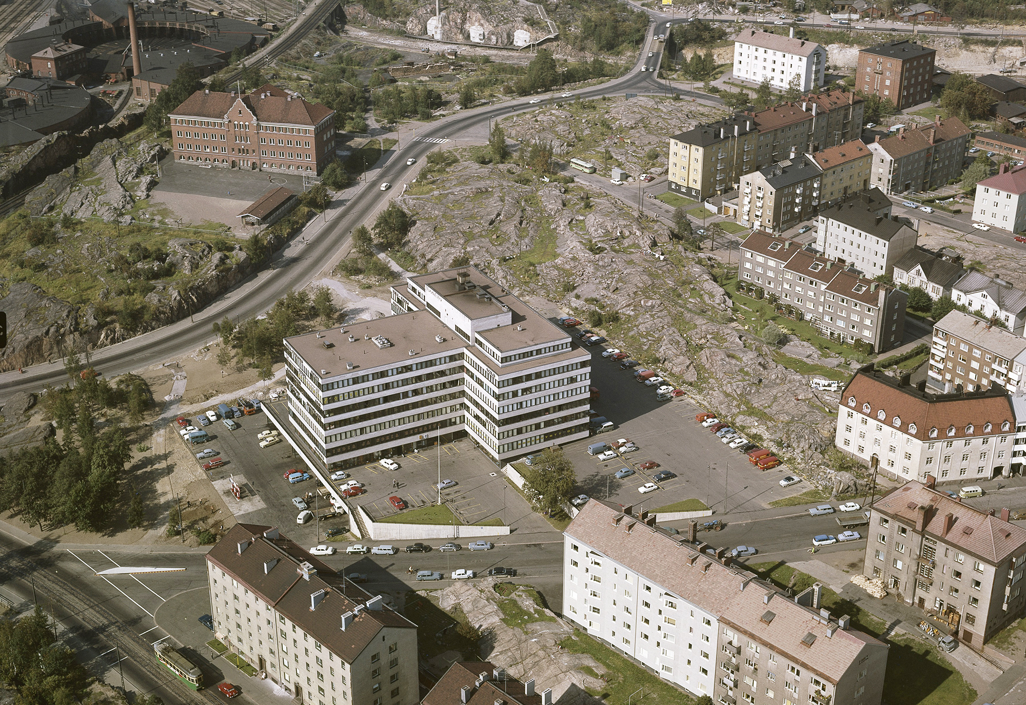 Kuvan keskellä T-kirjaimen muotoinen suuri modernistinen rakennus, ympärillä kalliota ja vanhempia kerrostaloja.