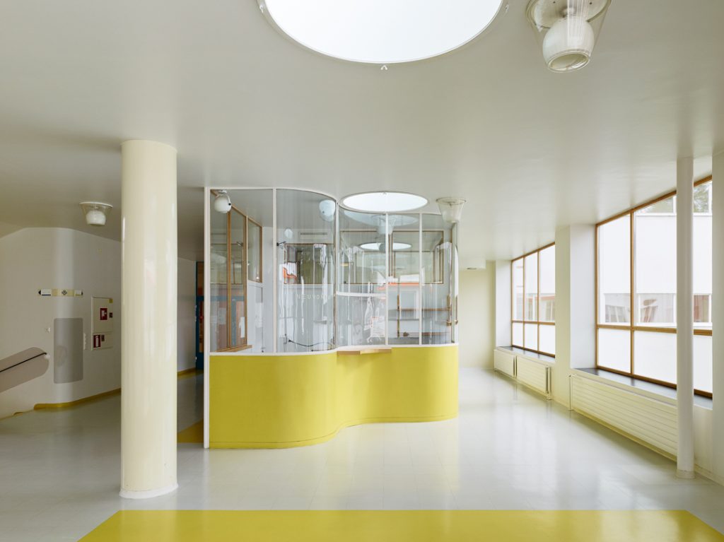 Paimion parantolan vastaanotto aula. Vastaanottokoppi on kaareileva ja sen ala osa on sitruunan keltainen, yläosa on lasia. 