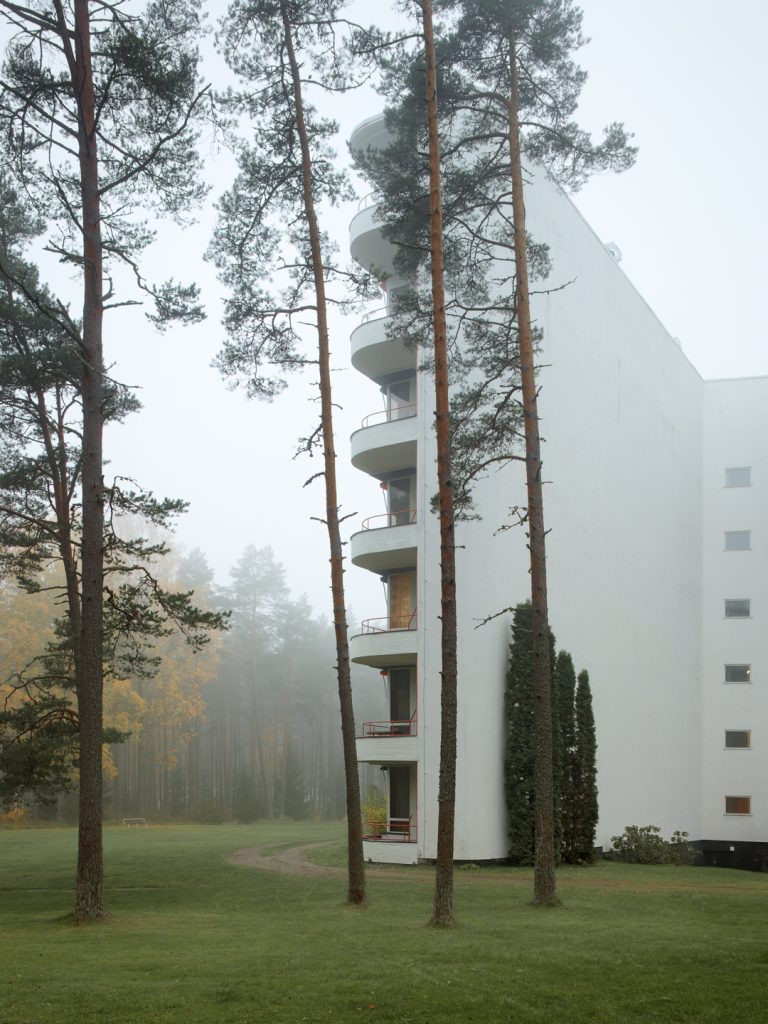 Paimio sanatorium's balcony wing's end seen from behind a few pine trees. The air is a little thick from the mist, but autumn foliaged trees shine through it.