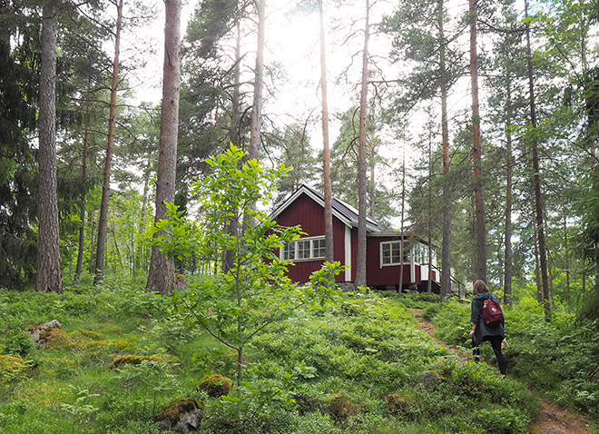 Punainen puinen puutalo keskellä metsää. Nainen kulkee polkua pitkin kohti taloa.