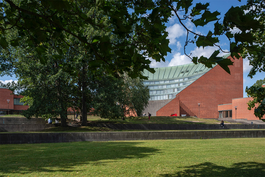 Aalto-yliopiston Kandidaattikeskuksen auditorio. Punatiilinen rakennus näkyy tuuheiden lehtipuiden takaa. Rakennuksen vihertävän kuparisen katon muoto muistuttaa amfiteatteria.
