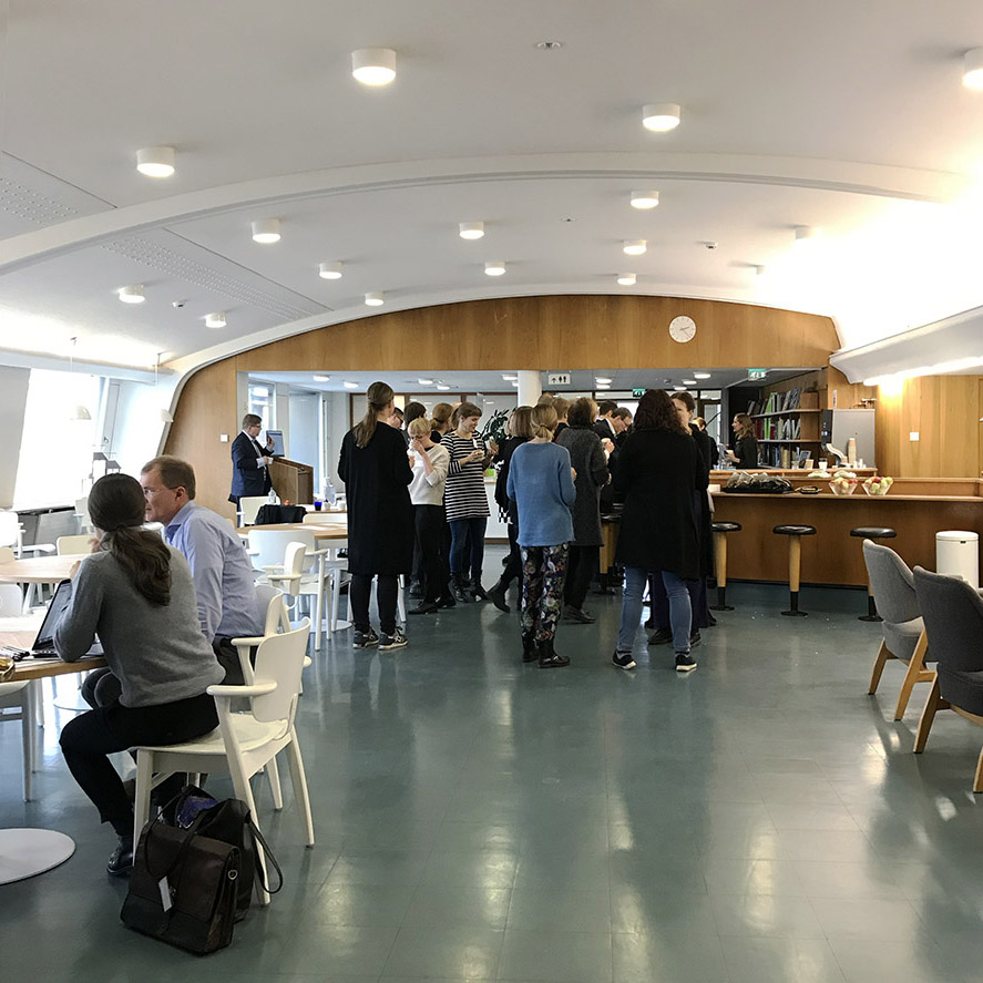 Voimatalon ylimmän kerroksen valoisassa aulatilassa vasemmalla pienryhmätyöskentelyä pöydän ääressä, taustalla ihmisryhmä kahvitauolla.