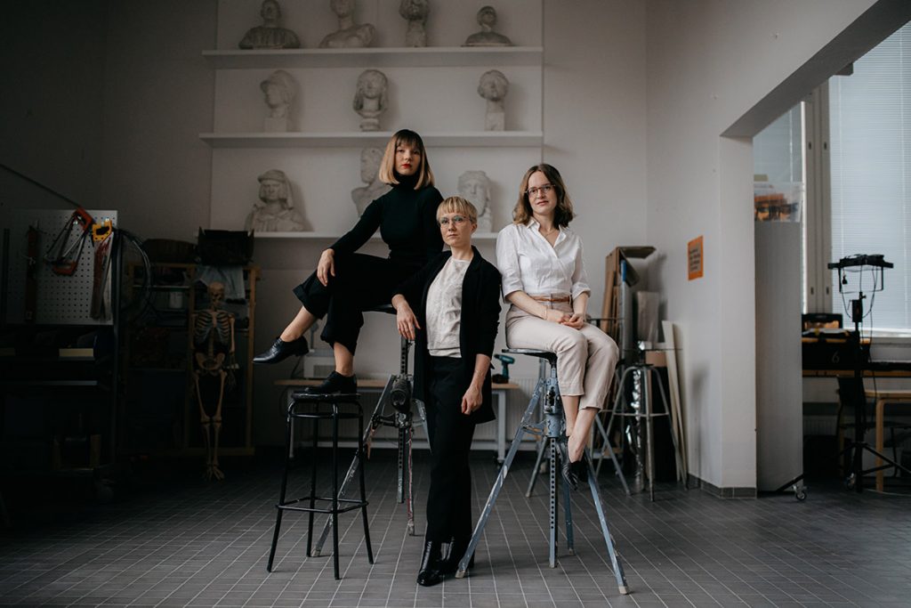 Kolme nuorta naista kuvanveistoateljeessa. Kaksi naisista istuu korkeilla statiiveilla, yksi seisoo. Korkean tilan takaseinällä hylly, jossa savesta tai kipsistä tehtyjä rintakuvaveistoksia.