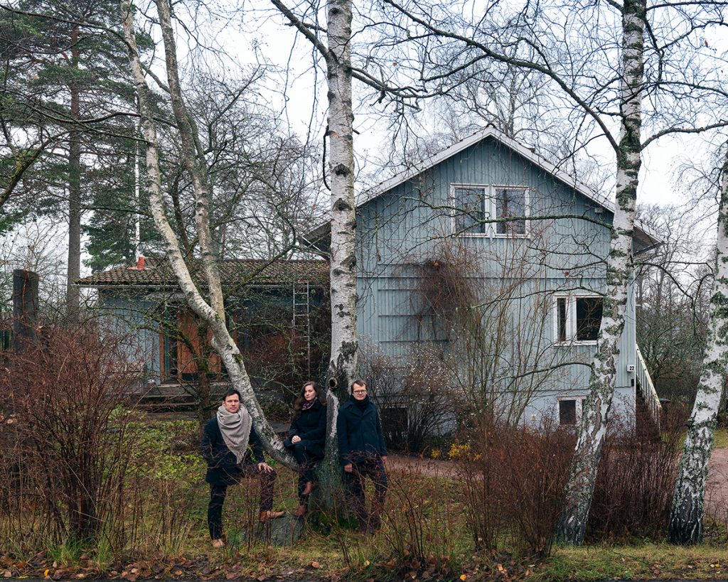 Three curators leaning agains birch trees in the front yard of a blue-gray wooden house.