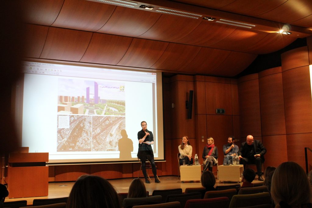 Five people in a panel discussion on stage of the Spaces of Peace Conference. Behind them a white board reflected with pictures of cities.