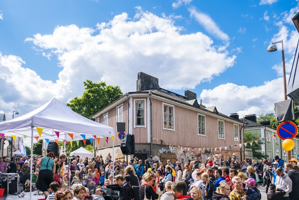 Helsingin Vallilan puutaloalueella katujuhlat aurinkoisena päivänä. Suuri joukko ihmisiä on kerääntynyt kadulle roosan värisen puutalon edustalle.