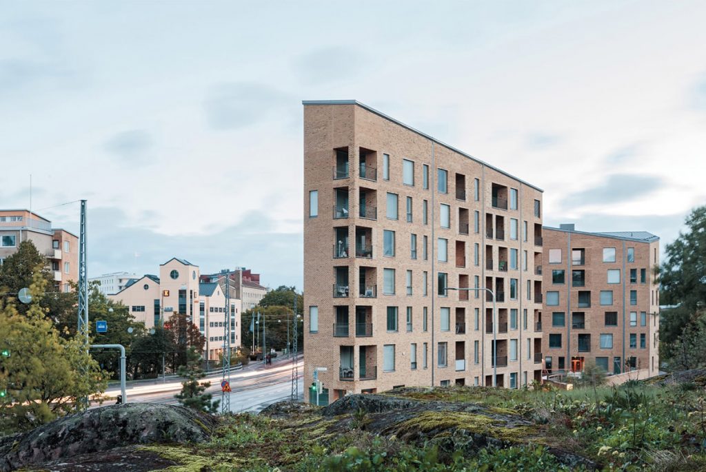 Narrow and wavy building along Mannerheimintie Street. The view opens from nearby rocks. The building façade is of burned red bricks and it has big windows.