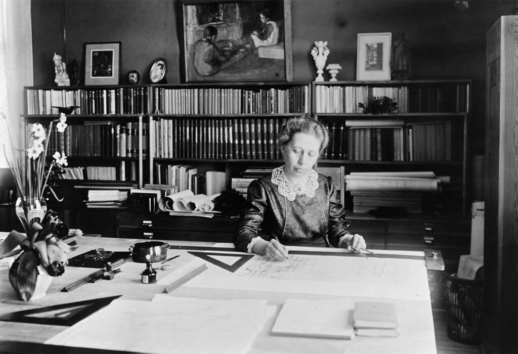 Wivi Lönn drawing a design by her desk in decorative 1910s clothes.