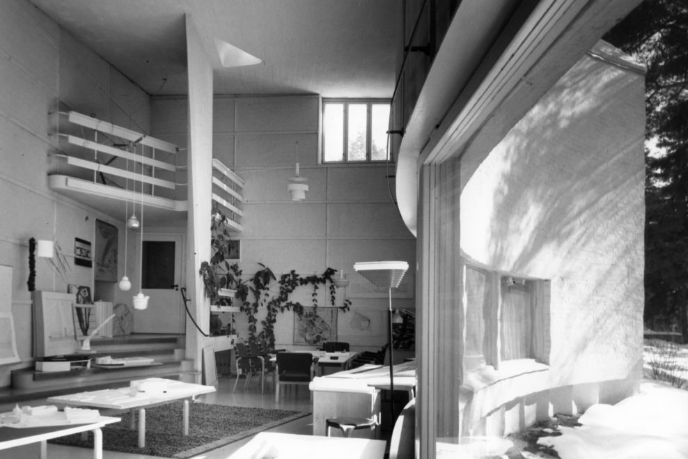Sunny view of Aalto's atelier. Sun shines in from ribbon windows on the curved wall. At the rear left are sloping stairs and above them a balcony-like structure with hanging lamps designed by Aalto. The plant climbs up along the wall of the room.