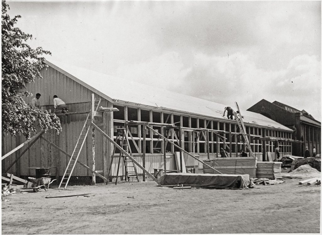 A wooden school under construction. Two men painton the facade, one man on the roof. Black and white.