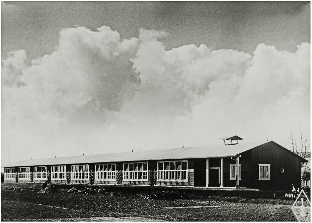 A long one-storey school building with a pitched roof. Dark-painted wooden facades, white window frames. Black and white.