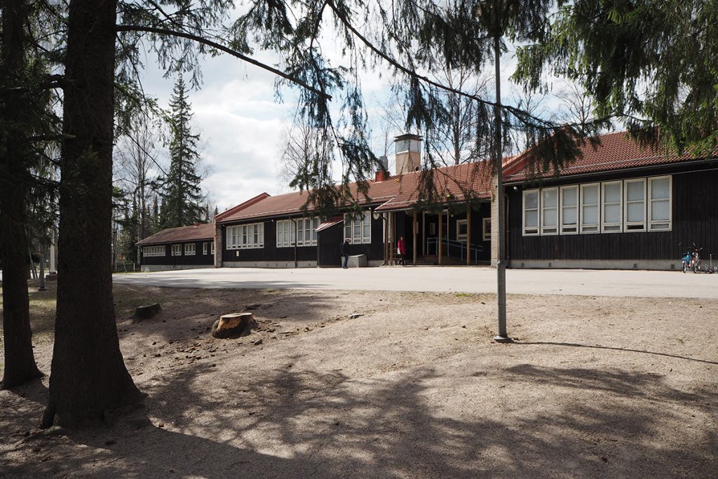 A view from under spruce tree branches towards the dark-painted school facade. Entrance canopy in the middle.