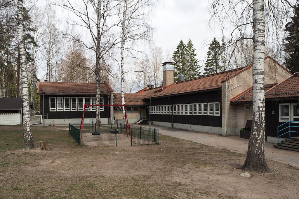 A playground with swings in the front, admist birch trees, and the dark-painted one-storey L-shaped school building in the background.