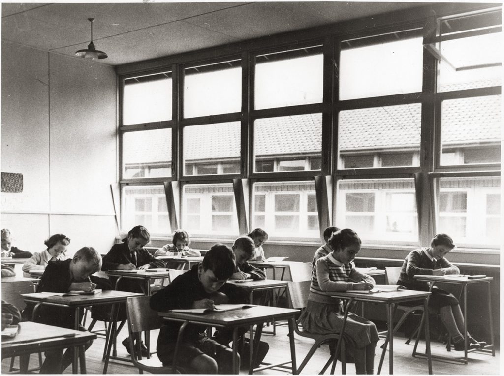 A classroom, children writing at their desks, a wall of windows in the background.