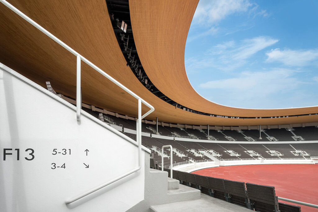 Wooden canopy curving over the stands. In the front, a white-rendered wall with section and row numbers.