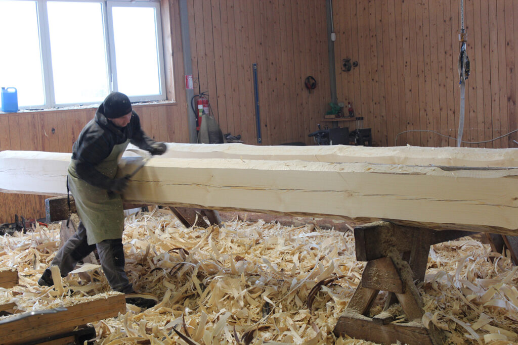 A man carves a big log in a workshop, the floor full of shavings.