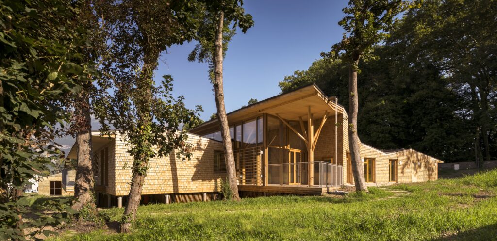 Moderni puinen talo vehreällä tontilla. Talo on tehty vaaleasta puusta ja sen muoto on epäsymmetrinen.