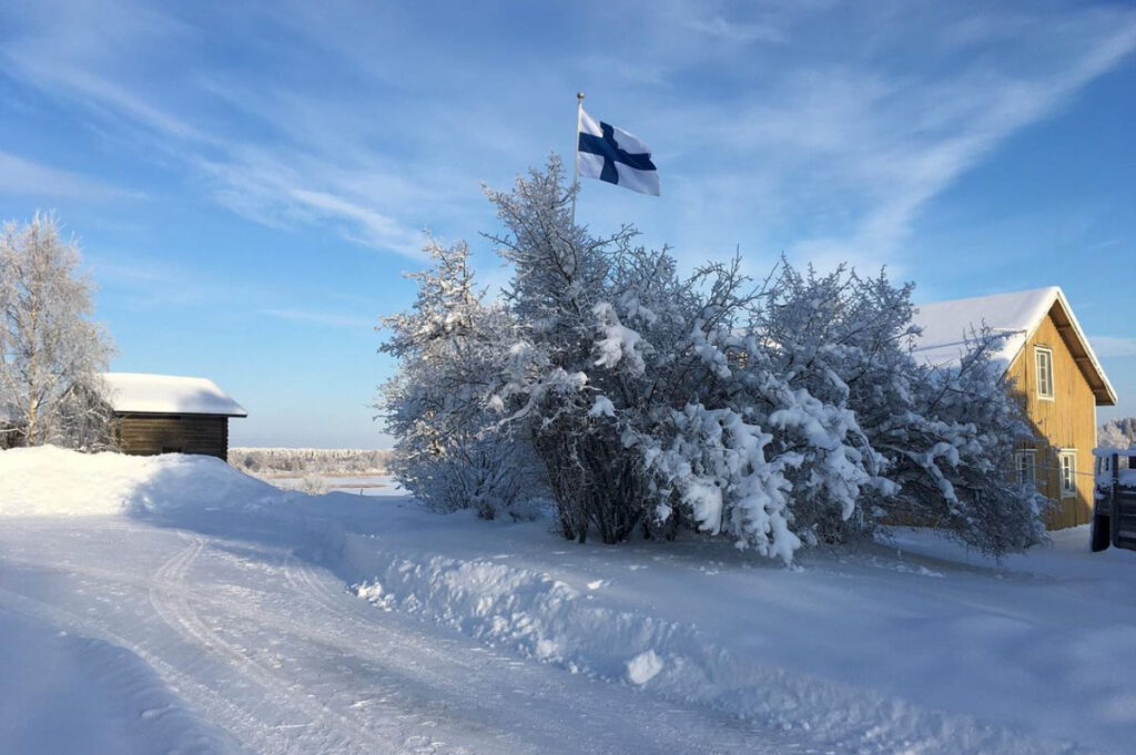 Suomen lippu nostettuna lipputankoon koulun pihassa aurinkoisen ja lumisen maiseman keskellä.