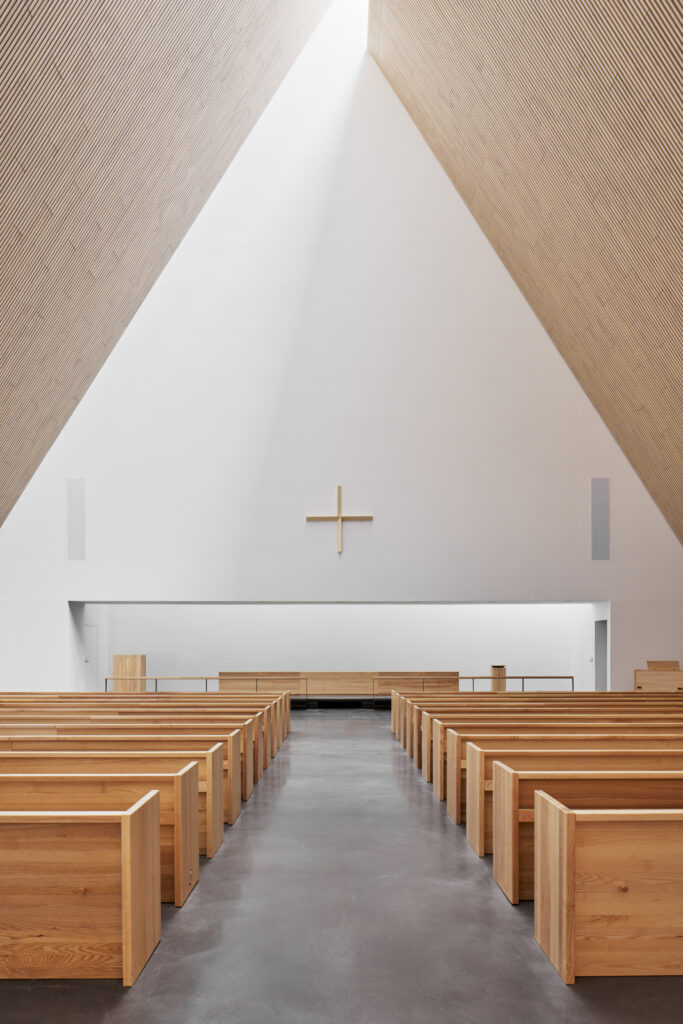 Frontal view towards altar, wooden benches and ceiling, white back wall with a delicate wooden cross