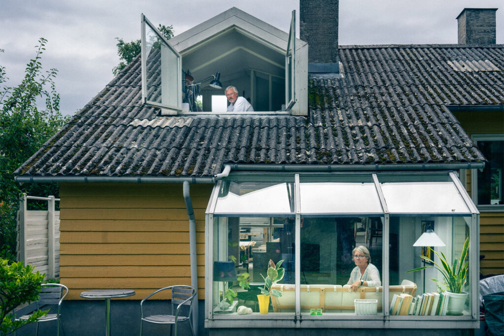 A yellow wooden house. A man looks outside from a window in the second floor. A woman stares to the viewer from inside a glassed-in veranda.