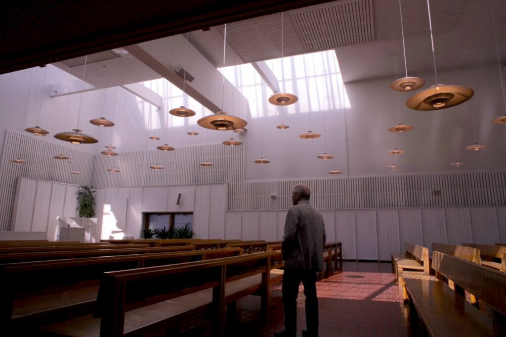 Man stands in a church hall, which has a red brick floor, white walls and indirect light from skylightes. A flock of lamps hanging half-way up.