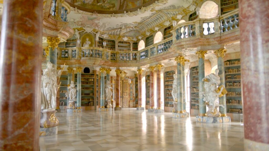 Rich Baroque interior with red and green marble columns, white statues, gilded decorations and bookshelves on along all walls. Light shines in through deep window niches.